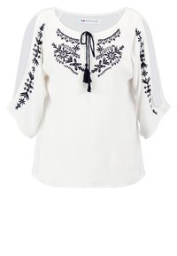 Blouse blanche avec des broderies florales bleu marine sur le devant. Coupe décontractée avec des manches trois-quarts et un lien à franges au cou.