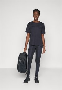 Sort træningstrøje med korte ærmer, parret med sorte mønstrede leggings og en mørk rygsæk. Modellen har sorte sneakers på.