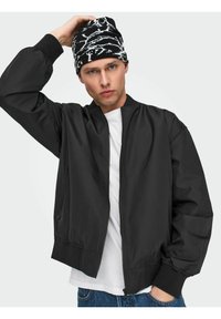 Black bomber jacket with a smooth texture and ribbed cuffs, worn over a white t-shirt. Accessory: black and white patterned knitted beanie.