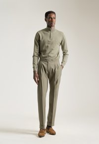 Jersey de cuello alto en verde olivo combinado con pantalones de talle alto y plisados en el mismo tono, y mocasines de ante marrón, todo ello sobre un fondo neutro.