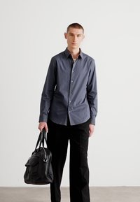 Μακρυμάνικη, πουκάμισο με κουμπιά σε σκούρο μπλε, συνδυασμένο με μαύρο παντελόνι. Η μοντέλο κρατά μια μαύρη δερμάτινη τσάντα. Απλό φόντο.