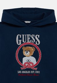 Μπλε ναυτικό φούτερ με μεγάλο κεντητό γραφικό αρκούδας σε κύκλο, διακοσμημένο με κόκκινο σχέδιο σχοινιού, με το όνομα της επωνυμίας "GUESS" από πάνω.