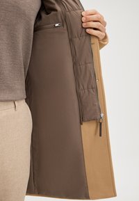 Cappotto esterno beige con una fodera interna liscia marrone; dispone di una tasca con zip e texture trapuntata all'interno.