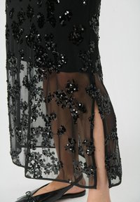 Robe noire avec superposition transparente, ornée de détails floraux en sequins. Fente à l'ourlet révélant la peau, portée avec des chaussures plates noires. Texture douce, design élégant.