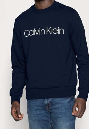 Mężczyzna ubrany w granatową bluzę z logo „Calvin Klein” na piersi oraz niebieskie dżinsy.