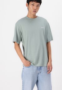 Tricou cu mânecă scurtă, de culoare verde deschis, dintr-un material moale, cu guler rotund și un logo mic "Club199" pe piept, purtat cu blugi din denim de culoare bleu deschis.