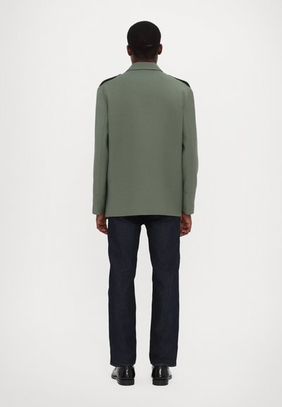 Manteau en mélange de laine vert à coupe décontractée et à manches longues, associé à un jean droit bleu foncé et des chaussures noires, vu de l'arrière.