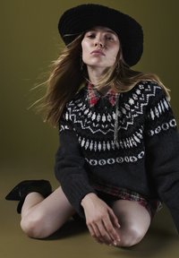 Maglione nero a maglia con motivi bianchi e grigi sopra una camicia a quadri rossa con colletto, indossato con un cappello nero a falda larga e stivali con tacco.