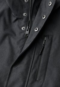 Veste noire en tissu texturé, dotée de fermetures éclair et de boutons-pression métalliques, comprenant une poche poitrine avec une fermeture éclair.