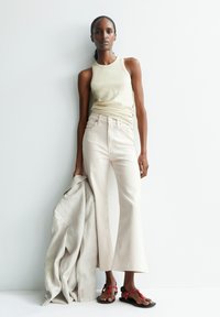 Lichtgroene geribbelde tanktop, lichtbeige high-waisted flared jeans, rode en zwarte sandalen, een beige linnen jack vasthoudend. Eenvoudig, modern ontwerp.