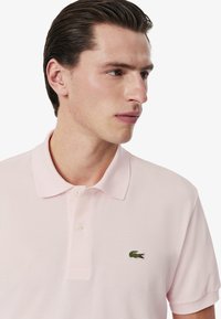 Ljusrosa polo shirt gjord av texturerad tyg, med en treknapps-knäppning och en liten broderad grön krokodil-logotyp på bröstet.