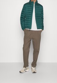 Teal puffer jacket with a zip front and high collar, worn over a white t-shirt. Paired with brown trousers and beige sneakers with black accents.