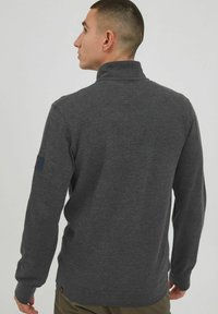 Gray, long-sleeve pullover in textured fabric with a high collar and logo patch on the left sleeve. Features a fitted silhouette.