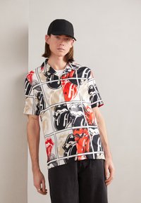 Camisa de botones de manga corta con un estampado gráfico de labios rojos, negros y Beige sobre un fondo blanco; tejido ligero; ajuste casual.