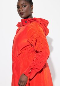 Veste rouge à capuche avec des coutures latérales froncées, fermeture éclair et pressions, manches longues et taille à cordon. Texture de tissu lisse et brillant.