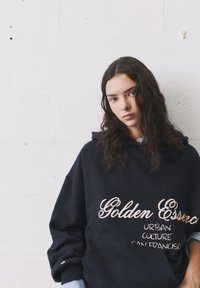 Námornícka oversized mikina s kapucňou s nápisom "Zlatá esencia" v kurzíve a "MESTSKÁ KULTÚRA SAN FRANCISCO" v tučnom písme. Bavlnený materiál s prednou vreckom.