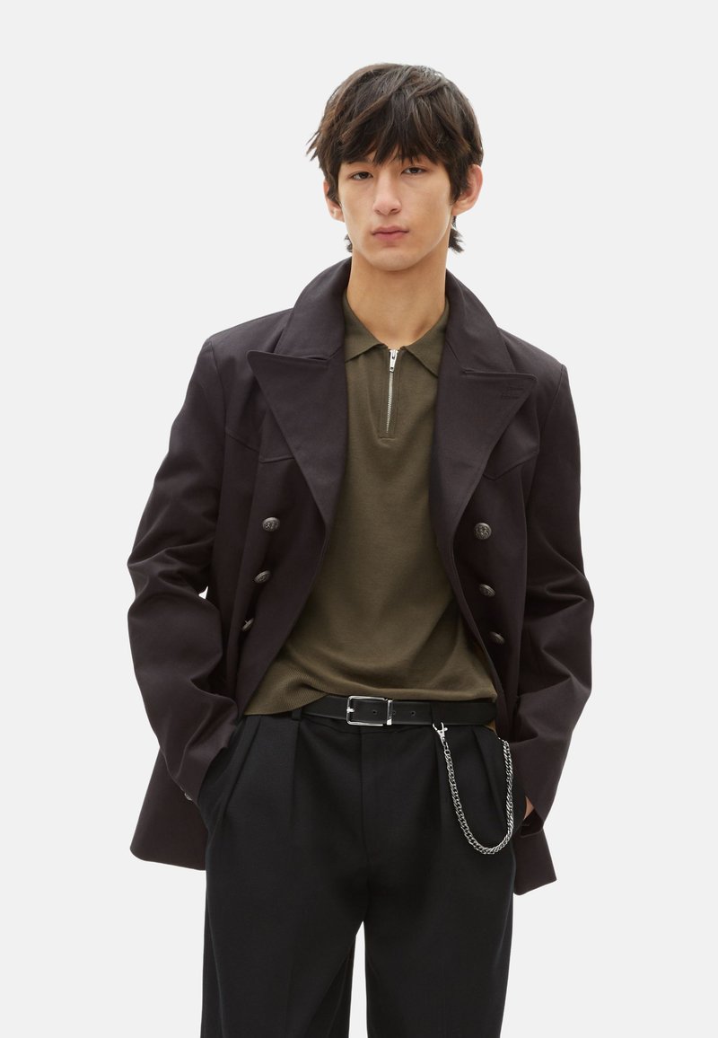 Schwarzer doppelt geknöpfter Mantel mit breiten Revers, olivgrünes Poloshirt, schwarze Hose mit Falten und silberner Kettendetail am Gürtel.