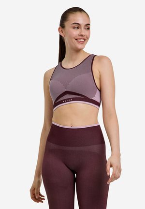 Junge Frau lächelt und schaut nach rechts, sie trägt ein passendes nahtloses lila und pinkes Sport-BH- und hoch tailliertes Leggings-Set.