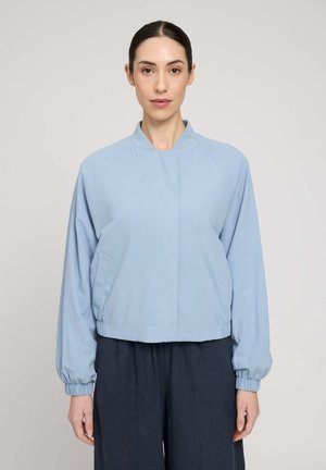 Woman with dark hair tied back wearing a light blue long-sleeve jacket and dark pants standing against a plain light background.
