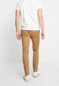 Pantaloni chino in tessuto beige con vestibilità slim, abbinati a una maglietta bianca semplice e scarpe da ginnastica bianche. Nessun motivo o decorazione visibile.