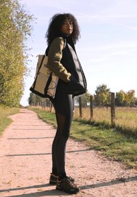 Fekete puffermellény egy zöld hosszúujjú felsőn, fekete leggings-szel, amelyen hálós panelek találhatók, és fekete túracipővel. Egy világos színű hátizsák.