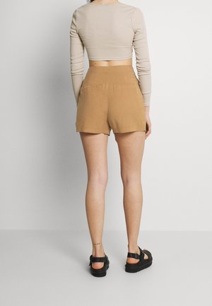 Kvinde stående med ryggen til iført en beige langærmet crop top, tanfarvede højtaljede shorts og sorte sandaler på en ensfarvet baggrund.
