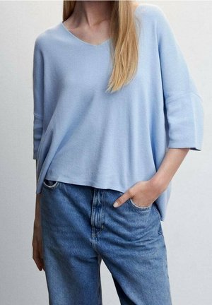 Nainen, joka pukeutuu vaaleansiniseen V-kaula-aukkoinen löysään neuleeseen ja korkeavyötäröisiin sinisiin farkkuihin, toinen käsi taskussa yksiväristä taustaa vasten.