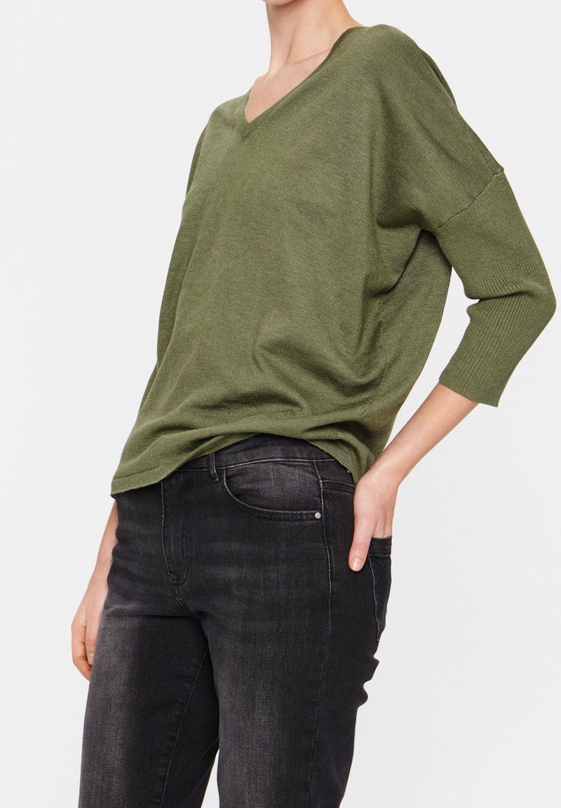 Personne portant un pull ample vert olive à col en V avec des manches trois-quarts et un jean noir délavé, la main reposant dans la poche arrière, sur fond blanc.