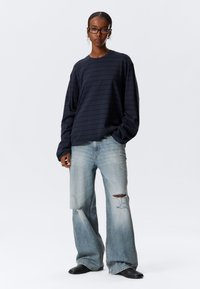 Μπλε ναυτικό ριγέ πουκάμισο με μακριά μανίκια, συνδυασμένο με ανοιχτό μπλε τζιν παντελόνι φαρδύ σε ίσια γραμμή με φθορές και μαύρα μποτάκια αστραγάλου, με άνετη εφαρμογή και υφή υφάσματος.