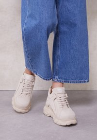 Fødder iført beige chunky sneakers og blå vidde bukser, der står på en grå gulv med en lys væg i baggrunden.