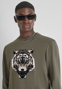 Sudadera de color verde oliva con un gráfico de tigre en blanco y negro y ojos amarillos. Modelo llevando unas gafas de sol negras y elegantes, mostrando una textura suave.