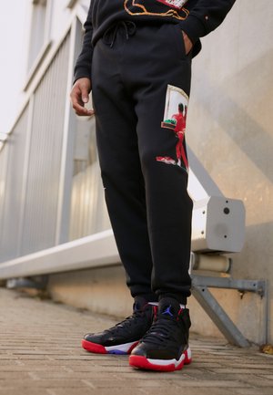 Persona vistiendo pantalones deportivos negros con un estampado de figura roja y zapatillas altas negras, rojas y azules, de pie en una acera pavimentada al aire libre.