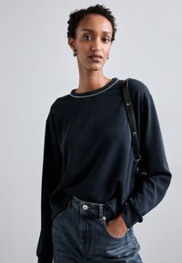 Top negro de manga larga con un cuello texturizado y decorado con cuentas. Combinado con unos vaqueros de tiro alto azules. La modelo sostiene un bolso de tiras negro. Tejido suave.