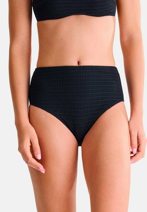 Femme portant un bas de bikini noir texturé taille haute et un haut assorti, debout les bras détendus le long du corps, sur un fond blanc uni.