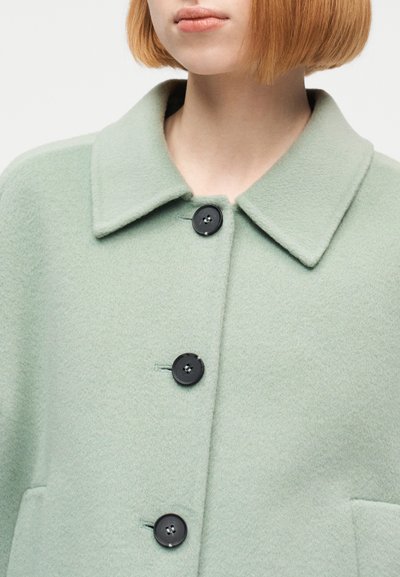 Γυναίκα με κοντό καρέ ξανθωπό-κόκκινο κούρεμα, φοράει παλτό από ανοιχτό πράσινο μάλλινο ύφασμα, κλεισμένο με τρία μεγάλα μ αύρα κουμπιά.