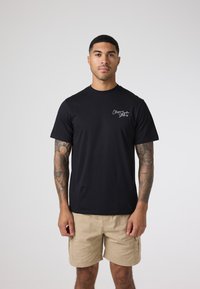 Czarny bawełniany t-shirt z krótkim rękawem i okrągłym dekoltem. Posiada małe białe logo po lewej stronie, noszony z beżowymi szortami cargo.