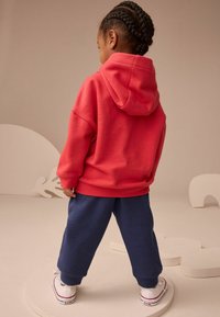 Bambino con capelli intrecciati che indossa un felpa rossa, pantaloni blu navy e sneakers bianche, in piedi su una piattaforma rotonda beige davanti a uno sfondo neutro.