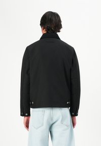 Black jacket featuring a classic collar, button cuffs, and snap closure details, paired with light blue jeans in a clean, minimalistic design.