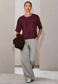 Mujer sosteniendo una bolsa marrón, vestida con una camiseta de manga corta burdeos, pantalones amplios grises y mocasines de color claro, caminando en el interior.