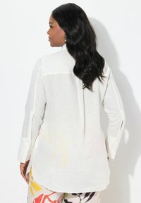 Witte linnen blouse met lange mouwen en zijslitsen, met een knoopmanschet. Van achter gezien, gecombineerd met kleurrijke, gedessineerde broek.