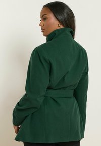 Vihreä, istuva takki, jossa on korkea kaulus ja vyötärölle sidottu vyö. Valmistettu teksturoidusta kankaasta, jossa on pitkät hihat ja pleatatut takaosat.