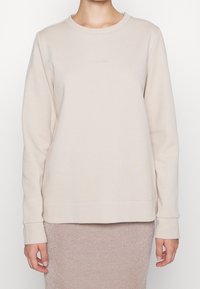 Persona che indossa un sweatshirt beige a maniche lunghe di Calvin Klein e una gonna rosa chiaro scintillante, mostrata dalla vita al collo.