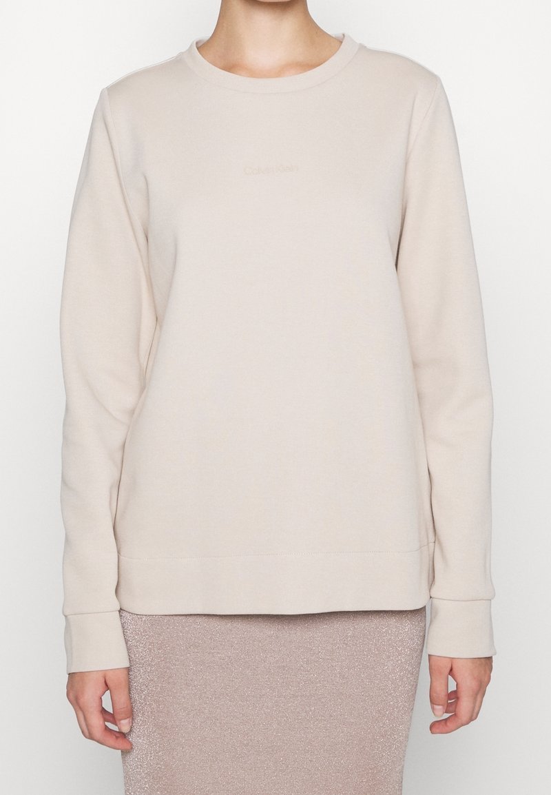 Persona che indossa un sweatshirt beige a maniche lunghe di Calvin Klein e una gonna rosa chiaro scintillante, mostrata dalla vita al collo.