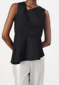 Top nero senza maniche con scollatura curva e orlo peplum, realizzato in un tessuto liscio. Indossato con pantaloni argentati lucenti.