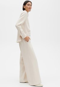 Roomkleurige op maat gemaakte blouse en broek met wijde pijpen van gladde stof. Heeft een kraag, manchetten met knopen en zakken.