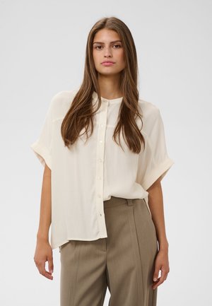 Femme aux longs cheveux bruns portant un chemisier ample crème boutonné et un pantalon beige taille haute, debout devant un fond uni.