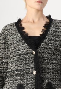 Svart-hvitt teksturert cardigan med frynser langs halslinjen, sølvknapper og V-formet krage.