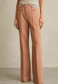 Pantalones a medida de color durazno con una textura suave, que cuentan con bolsillos laterales, pliegues marcados y una cintura ajustada con pasadores para cinturón.