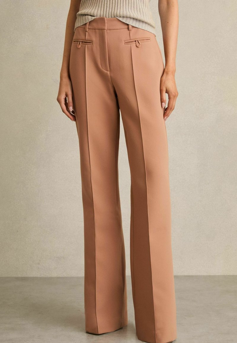 Pantalones a medida de color durazno con una textura suave, que cuentan con bolsillos laterales, pliegues marcados y una cintura ajustada con pasadores para cinturón.