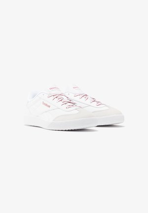 Par de zapatillas Reebok blancas de caña baja con marca y cordones en rosa pálido, suelas de goma y punteras de ante, inclinadas hacia la derecha sobre fondo blanco.